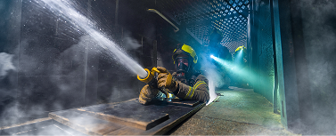 Firefighter sprays water through smoke-filled industrial corridor.
